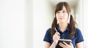紺色のユニフォーム(ポロシャツ)を着用した女性介護職員。廊下でメモ帳とペンを手に持ち、入居者の様子を思い浮かべるような真剣な表情で佇む様子。