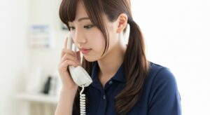 介護施設内で、若い女性介護職員が固定電話で利用者家族へ連絡している場面。表情は落ち着いており、状況報告や体調変化の共有、事故・ヒヤリハット後の経過説明などを行っている様子を示すイメージ。家族対応や情報共有の重要性を扱う文脈で使用可能。
