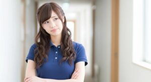 介護施設の廊下で腕を組み首をかしげる若い女性介護職員の様子。ケア方法や利用者対応について迷いながら考えている場面を想定したイメージで、認知症ケアや不穏対応、業務改善を検討する介護現場の課題を示す写真。