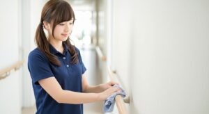 介護施設の廊下で、紺色のポロシャツを着た女性スタッフが手すりを丁寧に拭き掃除している様子