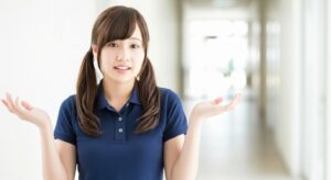 介護施設の廊下で、若い女性介護職員が両手を軽く広げている様子。状況説明や選択肢提示をしている場面、あるいは「どう対応すべきか」と考えながら周囲に問いかけているイメージ。