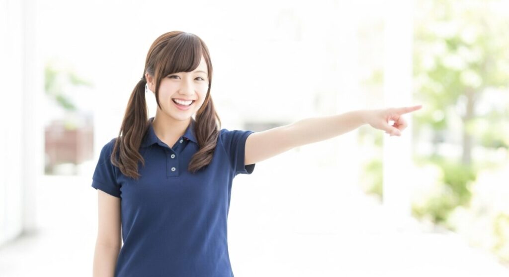 明るい室内で、若い女性介護職員が指を差しながら笑顔を見せている。案内や誘導をしている場面。