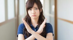 介護施設内で、若い女性介護職員が腕でバツ印を作り、困った表情を見せている。否定や注意を表現する場面。