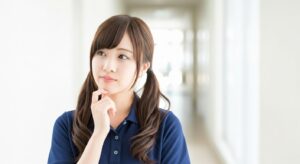 介護施設の廊下で、若い女性介護職員が顎に手を当てて考え込んでいる様子。ケア方法の選択や家族対応、記録の書き方などについて思案している場面を想起させるイメージ。