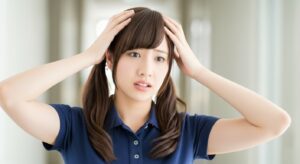 介護施設内で、ネイビーの制服を着た若い女性介護職員が両手を頭に当て、不安や困惑した表情を見せている様子。利用者対応や不穏症状への対応に悩み、精神的ストレスを抱えている介護現場の状況を表している場面。