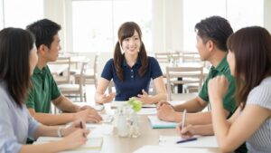 介護施設の明るい共有スペースで、ネイビーの制服を着た女性介護職員が中心となり、複数の職員とテーブルを囲んでミーティングを行っている様子。利用者ケアの方針や業務改善について意見交換し、チームケア体制を強化している場面。