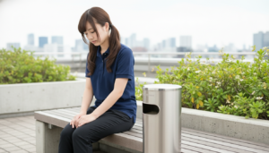 介護施設の屋上テラスで、ネイビーの制服を着た若い女性介護職員がベンチに座り、うつむいて手を膝に置きながら疲労した様子を見せている場面。