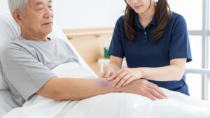 居室内で、若い女性介護職員が高齢男性の腕の内出血を確認している場面。転倒や打撲後の状態観察、皮下出血の有無や広がりをチェックしている状況を示すイメージ。