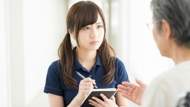 介護施設で、女性介護職員がメモ帳に記録を取りながら利用者の家族から話を聞いている様子。利用者の生活状況や要望を丁寧にヒアリングし、ケア内容の共有や情報収集を行っている場面。